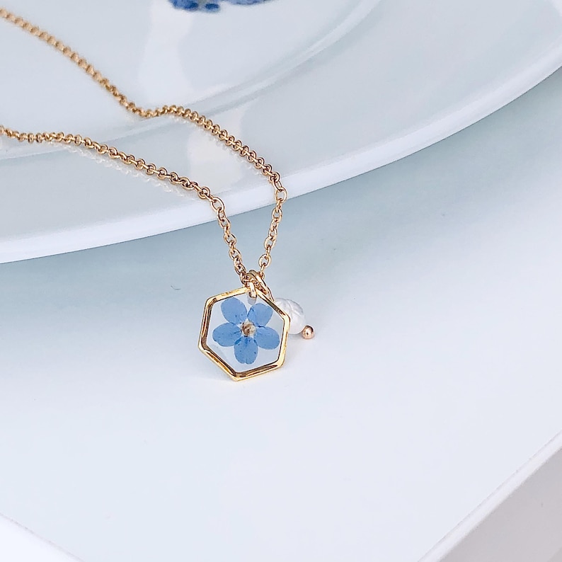 May include: A gold-colored necklace featuring a hexagonal pendant with a delicate blue flower encased within. The pendant is accented with a small white pearl. The necklace chain is also gold-colored, creating a cohesive and elegant design.