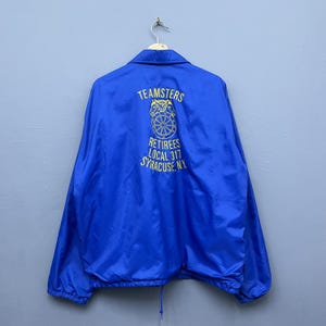 May include: A vibrant blue Teamsters jacket featuring yellow text and a logo on the back. The text reads "TEAMSTERS RETIREES LOCAL 317 SYRACUSE, NY". The jacket is suspended from a metal hanger.