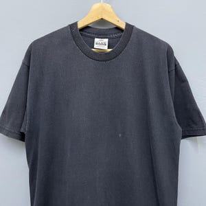 Op de afbeelding: Een zwart T-shirt met korte mouwen en een ronde hals, hangend aan een houten kleerhanger. Het shirt heeft een verweerde uitstraling en een klein wit label bij de halslijn. Het T-shirt is effen van kleur en lijkt van katoen te zijn.
