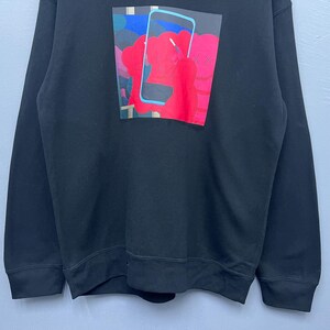 May include: A black long-sleeve sweatshirt featuring a rectangular graphic print on the chest. The print depicts a stylised red character with X-shaped eyes holding a light blue smartphone, set against a dark blue and purple background.
