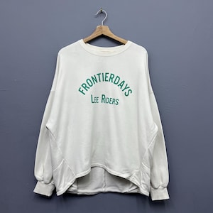 May include: Off-white sweatshirt with the text "FRONTIERDAYS" in green, arched above "Lee Riders". The long-sleeved top is hanging on a wooden hanger against a grey background. The sweatshirt has a rounded neckline and a slightly asymmetrical hemline.