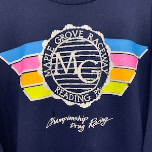 Może przedstawiać: Granatowa bluza z grafiką Maple Grove Raceway w Reading, PA. Wz&oacute;r zawiera okrągłe logo z literami MG, otoczone poziomymi paskami w kolorze niebieskim, r&oacute;żowym, pomarańczowym i ż&oacute;łtym, z napisem Championship Drag Racing poniżej.
