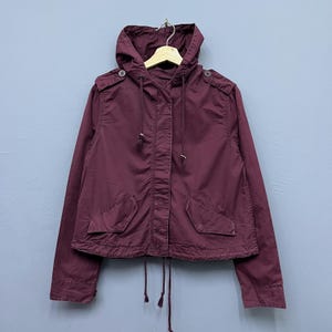 May include: A maroon hooded jacket with a drawstring closure and two front pockets. The jacket has long sleeves and is hanging on a wooden hanger. The jacket is made of a soft material and has a casual style.