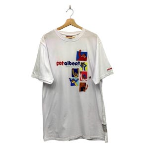 以下が含まれることがあります： 赤と青の刺繍で「Fat Albert」の文字が入った白いTシャツ。カラフルな漫画のキャラクターグラフィックが描かれています。半袖のシャツは木製のハンガーに掛けられています。