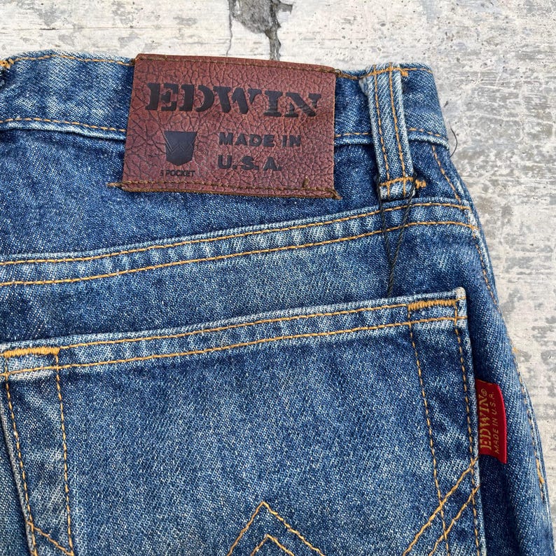Pode incluir: Close-up de jeans de ganga azul com costuras vis&iacute;veis e um remendo de couro castanho. O remendo diz "EDWIN MADE IN U.S.A." e &eacute; costurado na cintura. Uma etiqueta vermelha com "EDWIN MADE IN U.S.A." est&aacute; no bolso de tr&aacute;s.