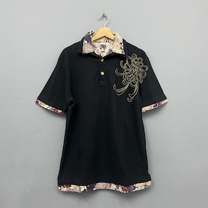 Puede incluir: Un polo negro de manga corta con un diseño de crisantemo bordado en dorado en el pecho. El cuello, los puños de las mangas y el dobladillo inferior están forrados con una tela de estampado floral multicolor. La prenda tiene tres botones.