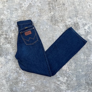 K&ouml;nnte beinhalten: Dunkelblaue Jeans aus Denim mit klassischem geradem Bein. Die Jeans hat einen braunen Lederaufn&auml;her mit dem Wrangler-Logo auf der Ges&auml;&szlig;tasche und ein kleines goldenes Etikett &uuml;ber der Ges&auml;&szlig;tasche. Die Jeans liegt flach auf einer grauen Oberfl&auml;che.
