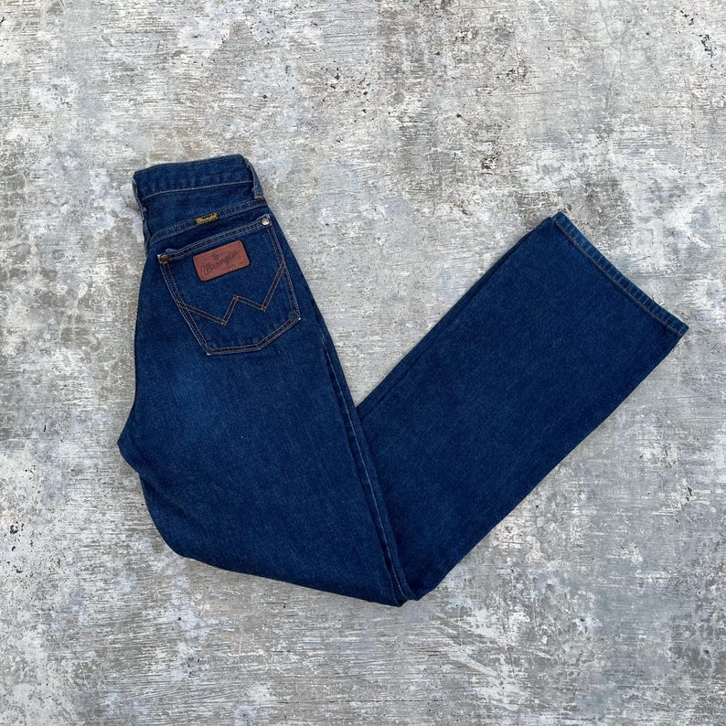 K&ouml;nnte beinhalten: Dunkelblaue Jeans aus Denim mit klassischem geradem Bein. Die Jeans hat einen braunen Lederaufn&auml;her mit dem Wrangler-Logo auf der Ges&auml;&szlig;tasche und ein kleines goldenes Etikett &uuml;ber der Ges&auml;&szlig;tasche. Die Jeans liegt flach auf einer grauen Oberfl&auml;che.