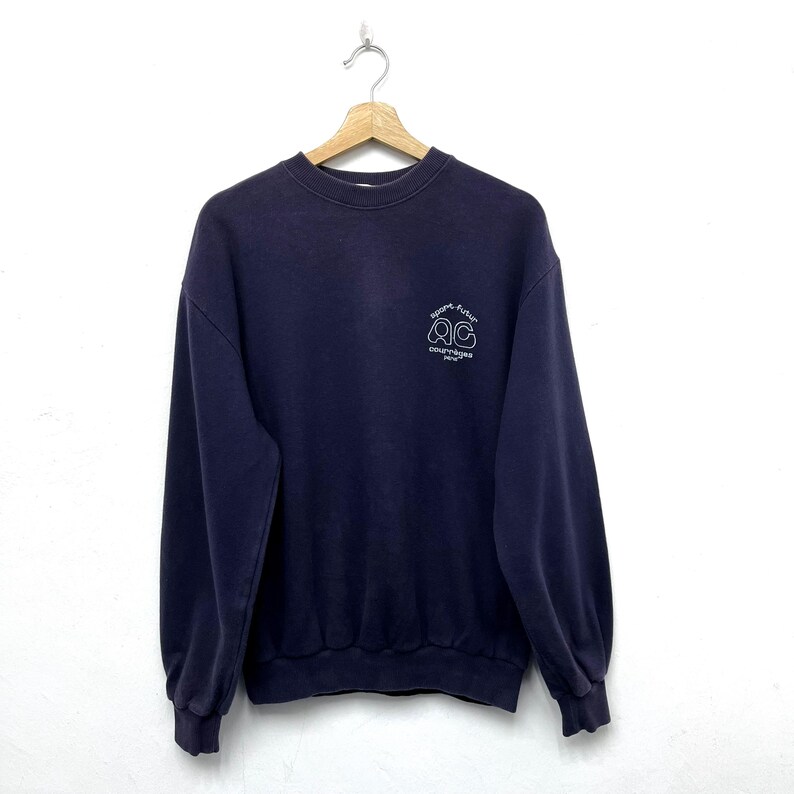 May include: A navy blue sweatshirt with a crew neck and long sleeves. The sweatshirt features a small white graphic on the upper left chest that reads "sport, future, courreges, Paris". The garment is hanging from a metal hanger.