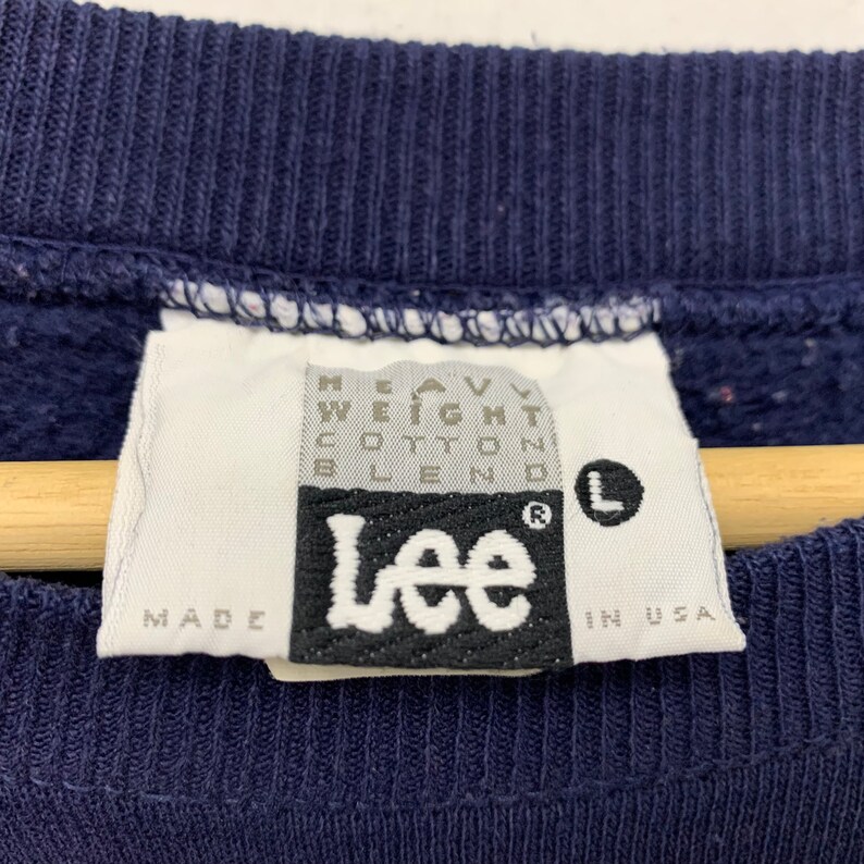 Może przedstawiać: Granatowa bluza z białym materiałowym metką z napisem HEAVY WEIGHT COTTON BLEND, logo Lee oraz literą L w czarnym k&oacute;łku. Metka zawiera r&oacute;wnież napis MADE IN USA, wskazujący na kraj pochodzenia produktu.