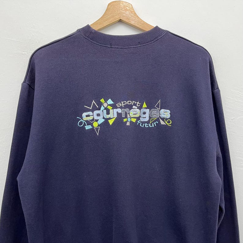 May include: Navy blue sweatshirt with the text "courr&egrave;ges sport futur" in a colorful, geometric design. The sweatshirt is hanging on a wooden hanger.