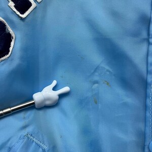 May include: Light blue jacket with a white and navy blue patch. The jacket has a button closure and a white hand-shaped pointer. The jacket has some stains.
