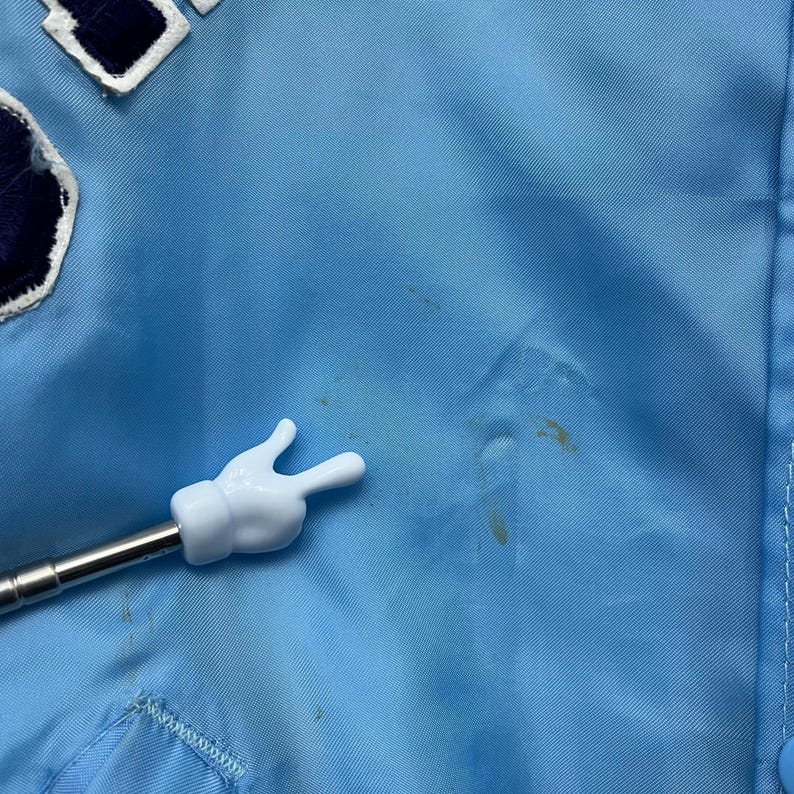 May include: Light blue jacket with a white and navy blue patch. The jacket has a button closure and a white hand-shaped pointer. The jacket has some stains.