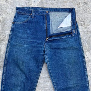 Pode incluir: Close-up de cal&ccedil;as de ganga azuis com uma braguilha parcialmente aberta. As cal&ccedil;as de ganga t&ecirc;m um design cl&aacute;ssico de cinco bolsos, com fecho de bot&atilde;o e fecho de correr. O denim tem um aspeto ligeiramente desbotado.