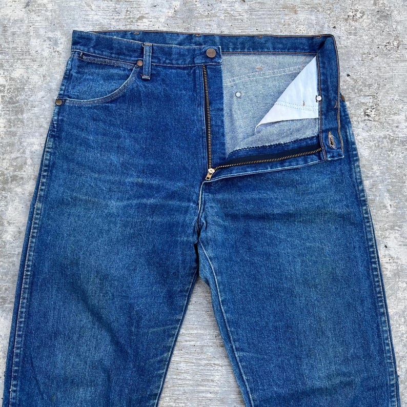Pode incluir: Close-up de cal&ccedil;as de ganga azuis com uma braguilha parcialmente aberta. As cal&ccedil;as de ganga t&ecirc;m um design cl&aacute;ssico de cinco bolsos, com fecho de bot&atilde;o e fecho de correr. O denim tem um aspeto ligeiramente desbotado.