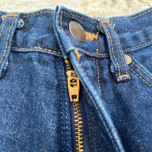 K&ouml;nnte beinhalten: Nahaufnahme einer blauen Jeans aus Denim mit einem Messingrei&szlig;verschluss und einem Knopfverschluss. Die Jeans hat orangefarbene N&auml;hte und einen Metallknopf mit dem Markennamen. Der Rei&szlig;verschluss ist teilweise ge&ouml;ffnet.