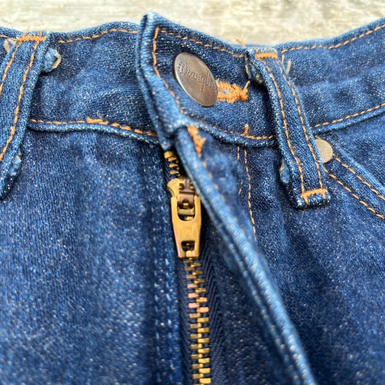 K&ouml;nnte beinhalten: Nahaufnahme einer blauen Jeans aus Denim mit einem Messingrei&szlig;verschluss und einem Knopfverschluss. Die Jeans hat orangefarbene N&auml;hte und einen Metallknopf mit dem Markennamen. Der Rei&szlig;verschluss ist teilweise ge&ouml;ffnet.