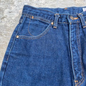 K&ouml;nnte beinhalten: Nahaufnahme einer blauen Jeans. Die Jeans hat ein klassisches F&uuml;nf-Taschen-Design mit orangefarbenen N&auml;hten und Metallnieten. Der Stoff ist dunkel gewaschen und die Jeans scheinen Vintage zu sein.
