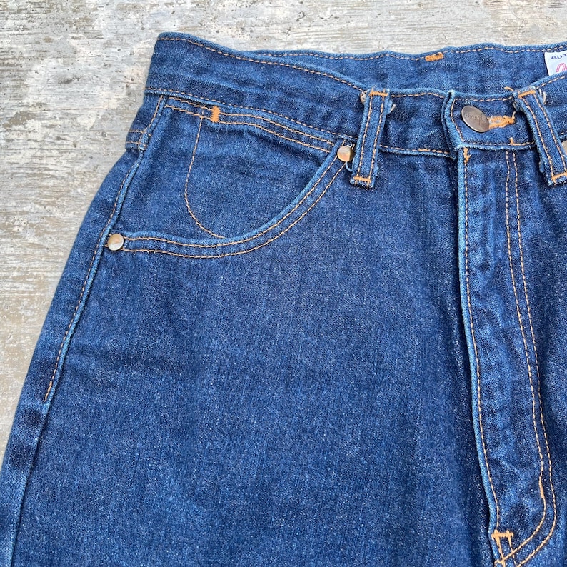 K&ouml;nnte beinhalten: Nahaufnahme einer blauen Jeans. Die Jeans hat ein klassisches F&uuml;nf-Taschen-Design mit orangefarbenen N&auml;hten und Metallnieten. Der Stoff ist dunkel gewaschen und die Jeans scheinen Vintage zu sein.