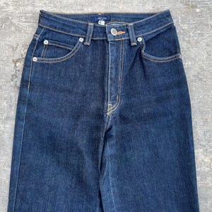 May include: Dark wash denim jeans with a high waist and straight leg. The jeans feature classic five-pocket styling, a button and zipper closure, and contrasting yellow stitching. The jeans are a size 6.