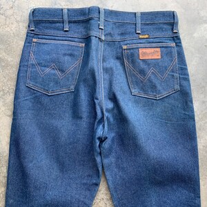 K&ouml;nnte beinhalten: Dunkelblaue Jeans aus Denim mit klassischem F&uuml;nf-Taschen-Design. Merkmale sind ein brauner Lederaufn&auml;her mit dem "Wrangler"-Logo und charakteristische N&auml;hte auf den Ges&auml;&szlig;taschen. Die Jeans hat einen geraden Beinverlauf und einen Knopf- und Rei&szlig;verschluss.