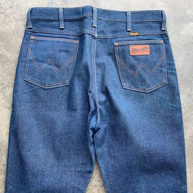K&ouml;nnte beinhalten: Dunkelblaue Jeans aus Denim mit klassischem F&uuml;nf-Taschen-Design. Merkmale sind ein brauner Lederaufn&auml;her mit dem "Wrangler"-Logo und charakteristische N&auml;hte auf den Ges&auml;&szlig;taschen. Die Jeans hat einen geraden Beinverlauf und einen Knopf- und Rei&szlig;verschluss.