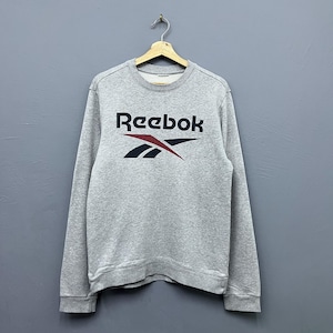 Op de afbeelding: Een grijze sweatshirt met het Reebok-logo in zwart, rood en marineblauw. De sweatshirt hangt aan een houten hanger tegen een grijze achtergrond. Het Reebok-logo staat in het midden op de borst.
