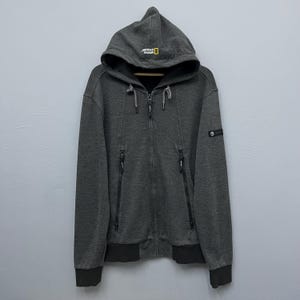 以下が含まれることがあります： チャコールグレーのジップアップパーカー。フードと2つのフロントポケットが付いています。フードには「National Geographic」のロゴ、袖には小さなエンブレムが付いています。袖口と裾は濃いグレーです。
