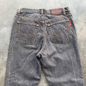 May include: Back view of gray denim jeans with a classic five-pocket design. The jeans feature a brown leather patch at the waistband and a red logo tag. The back pockets have a distinctive stitched design in a contrasting tan color.