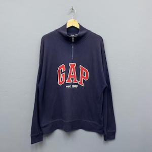 May include: Navy blue sweatshirt with a mock neck and a quarter-zip. The red "GAP" logo with a white outline and "est. 1969" is printed on the front. The sweatshirt is suspended from a wooden hanger.