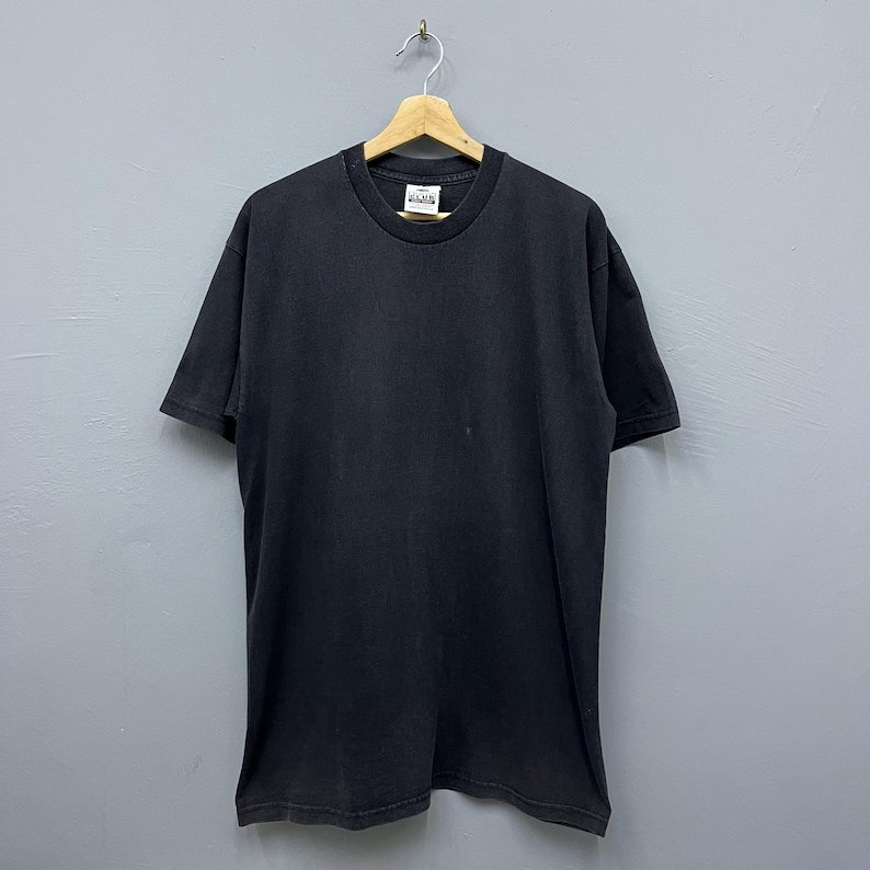 Op de afbeelding: Een zwart T-shirt met korte mouwen, hangend aan een houten hanger. Het shirt met ronde hals heeft een licht vervaagde uitstraling. Een label is zichtbaar bij de halslijn. De achtergrond is een lichtgrijze muur.
