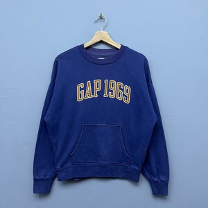 Könnte beinhalten: Ein marineblaues Sweatshirt mit Kängurutasche und dem Text "GAP 1969" in goldfarbenen Buchstaben. Das Sweatshirt hängt an einem Holzbügel vor einem hellblauen Hintergrund. Das Kleidungsstück hat einen Vintage-Look.