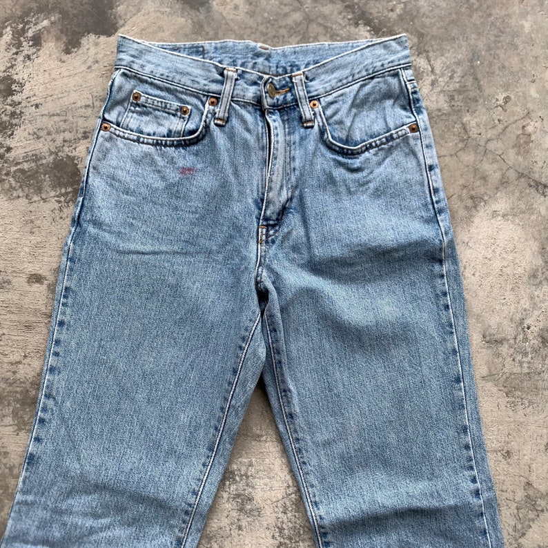 K&ouml;nnte beinhalten: Hellblaue Jeans mit klassischem F&uuml;nf-Taschen-Design. Die Jeans haben einen Knopf- und Rei&szlig;verschluss, kupferfarbene Nieten und einen kleinen roten Fleck in der N&auml;he der Vordertasche. Vintage-Stil.