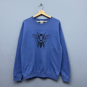 Puede incluir: Sudadera azul con un gráfico de abeja negra. La sudadera de cuello redondo está colgada de una percha de madera sobre un fondo gris. El gráfico de la abeja está centrado en la parte delantera de la sudadera.