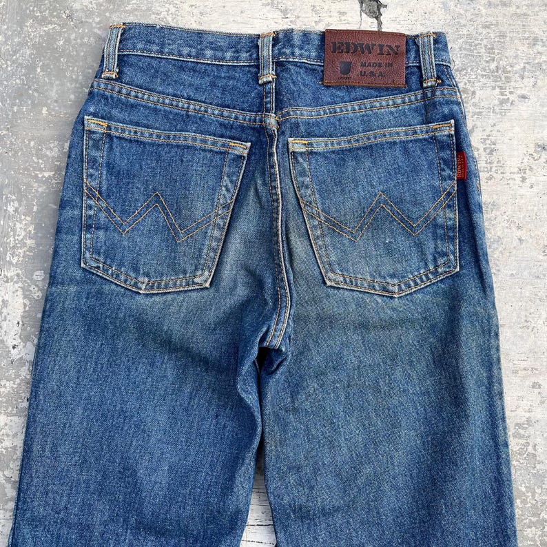 Pode incluir: Jeans azuis de ganga com um design cl&aacute;ssico de cinco bolsos. Os bolsos traseiros apresentam um padr&atilde;o de costura distinto. Uma etiqueta de couro castanho com a marca "EDWIN" &eacute; vis&iacute;vel. Os jeans s&atilde;o feitos nos EUA.