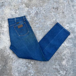 May include: Blue denim jeans with a classic straight leg cut. The jeans feature a brown leather patch on the back pocket with the brand logo. The jeans are folded neatly on a gray concrete surface.