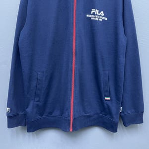 Puede incluir: Chaqueta azul marino con cremallera, una raya roja en el centro y el logotipo blanco de FILA en el pecho. La chaqueta tiene dos bolsillos delanteros y un peque&ntilde;o logotipo en la manga izquierda. El texto "MODA NELLA VITA SPORTIVA NUMERO UNO" est&aacute; impreso debajo del logotipo de FILA.
