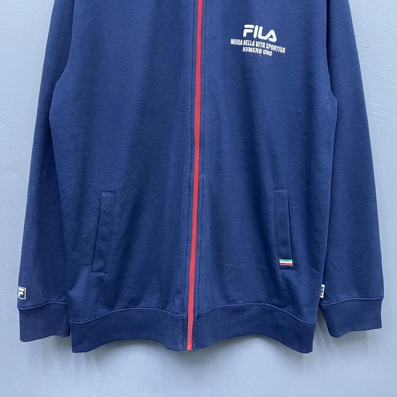 Puede incluir: Chaqueta azul marino con cremallera, una raya roja en el centro y el logotipo blanco de FILA en el pecho. La chaqueta tiene dos bolsillos delanteros y un peque&ntilde;o logotipo en la manga izquierda. El texto "MODA NELLA VITA SPORTIVA NUMERO UNO" est&aacute; impreso debajo del logotipo de FILA.
