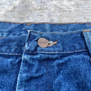 Pode incluir: Close-up de jeans azuis com um bot&atilde;o cor de cobre. O bot&atilde;o &eacute; gravado com a palavra "Wrangler". Os jeans t&ecirc;m costuras laranja.