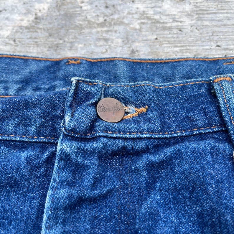 Pode incluir: Close-up de jeans azuis com um bot&atilde;o cor de cobre. O bot&atilde;o &eacute; gravado com a palavra "Wrangler". Os jeans t&ecirc;m costuras laranja.