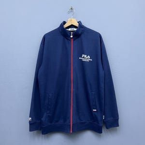 Puede incluir: Chaqueta azul marino con cremallera y una raya roja en la cremallera. La chaqueta presenta el nombre de la marca "FILA" en blanco en el pecho y un peque&ntilde;o logotipo en la manga. Tiene dos bolsillos delanteros y cuello alto.