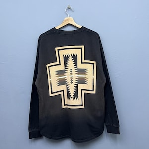 Könnte beinhalten: Schwarzes Langarm-Sweatshirt mit einem großen Kreuzdesign auf dem Rücken. Das Kreuz hat einen beigen Rand und ein zentrales Muster mit Zickzack- und geometrischen Formen in Braun-, Grün- und Beigetönen. Das Sweatshirt hängt an einem Holzbügel.
