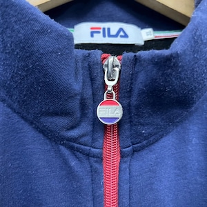 Puede incluir: Primer plano de una sudadera con cremallera azul marino. La cremallera roja tiene un dije circular con el nombre de la marca "FILA" en blanco. El cuello tiene una raya blanca, verde y roja. El nombre de la marca "FILA" tambi&eacute;n es visible.