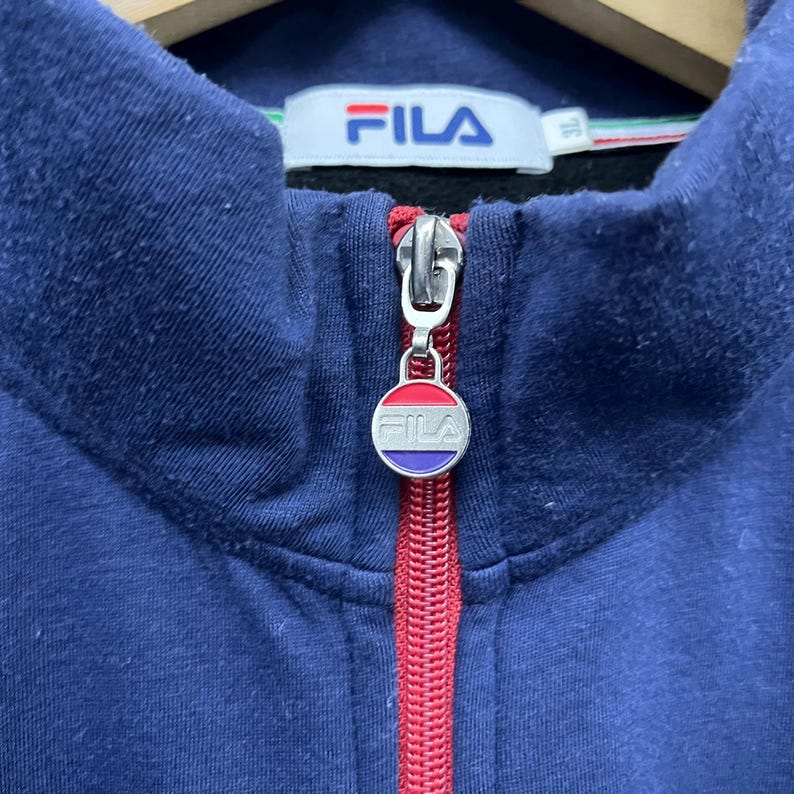 Puede incluir: Primer plano de una sudadera con cremallera azul marino. La cremallera roja tiene un dije circular con el nombre de la marca "FILA" en blanco. El cuello tiene una raya blanca, verde y roja. El nombre de la marca "FILA" tambi&eacute;n es visible.