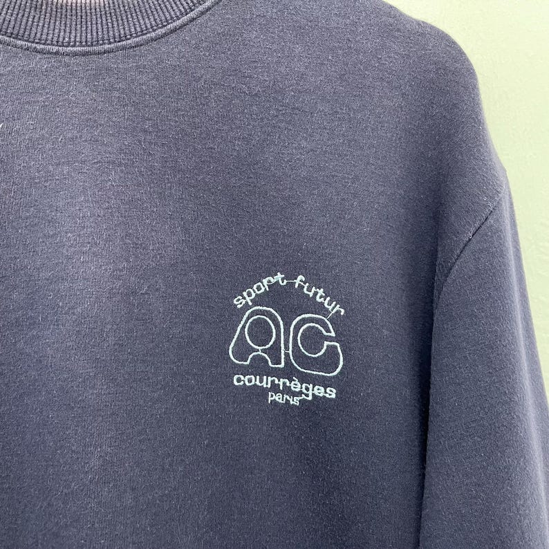 May include: A navy blue sweatshirt with a crew neck. The white text on the front reads "sport futur" above the letters "AC" and the word "courr&egrave;ges paris". The sweatshirt is made of a soft material.