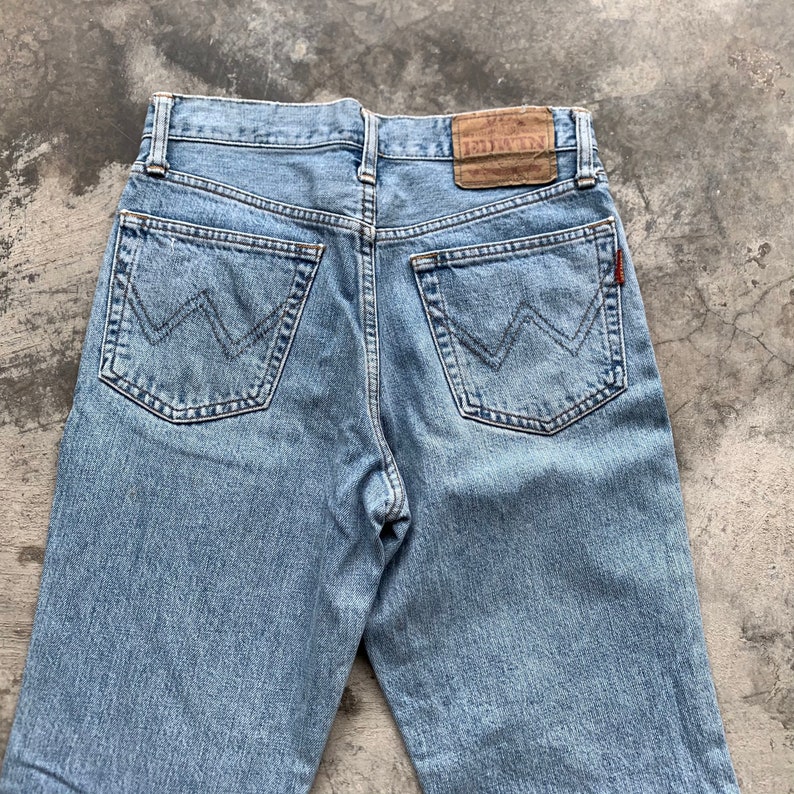 K&ouml;nnte beinhalten: Hellblaue Jeans mit dem Markennamen "EDWIN" auf einem braunen Lederpatch. Die Jeans hat zwei Ges&auml;&szlig;taschen mit einem markanten "W"-Nahtdesign. Ein kleines rotes Etikett ist auf der rechten Ges&auml;&szlig;tasche sichtbar.
