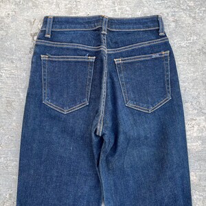 May include: Dark wash denim jeans with a high waist and two back pockets. The jeans have a classic five-pocket design with a straight leg. The stitching is a contrasting light brown color. The jeans are a vintage style.