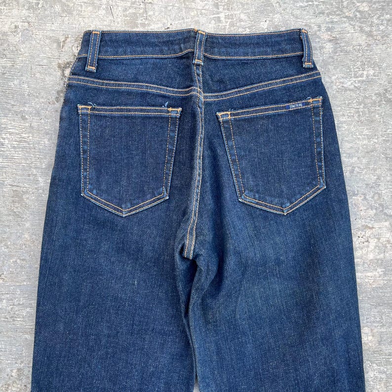 May include: Dark wash denim jeans with a high waist and two back pockets. The jeans have a classic five-pocket design with a straight leg. The stitching is a contrasting light brown color. The jeans are a vintage style.
