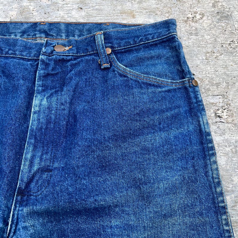 Pode incluir: Close-up de jeans de ganga azul. Os jeans t&ecirc;m um design cl&aacute;ssico de cinco bolsos com fecho de bot&atilde;o e z&iacute;per. O denim tem uma apar&ecirc;ncia ligeiramente desbotada, com desgaste vis&iacute;vel.