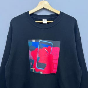 May include: A black crewneck sweatshirt featuring a graphic print on the chest. The design depicts a bright red cartoon character with X-shaped eyes holding a light blue smartphone. The garment has a ribbed neckline and a white interior brand label.